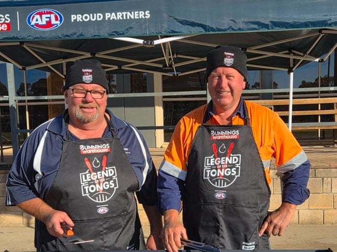 Two men holding tongs standing behind a Barbecue outside.