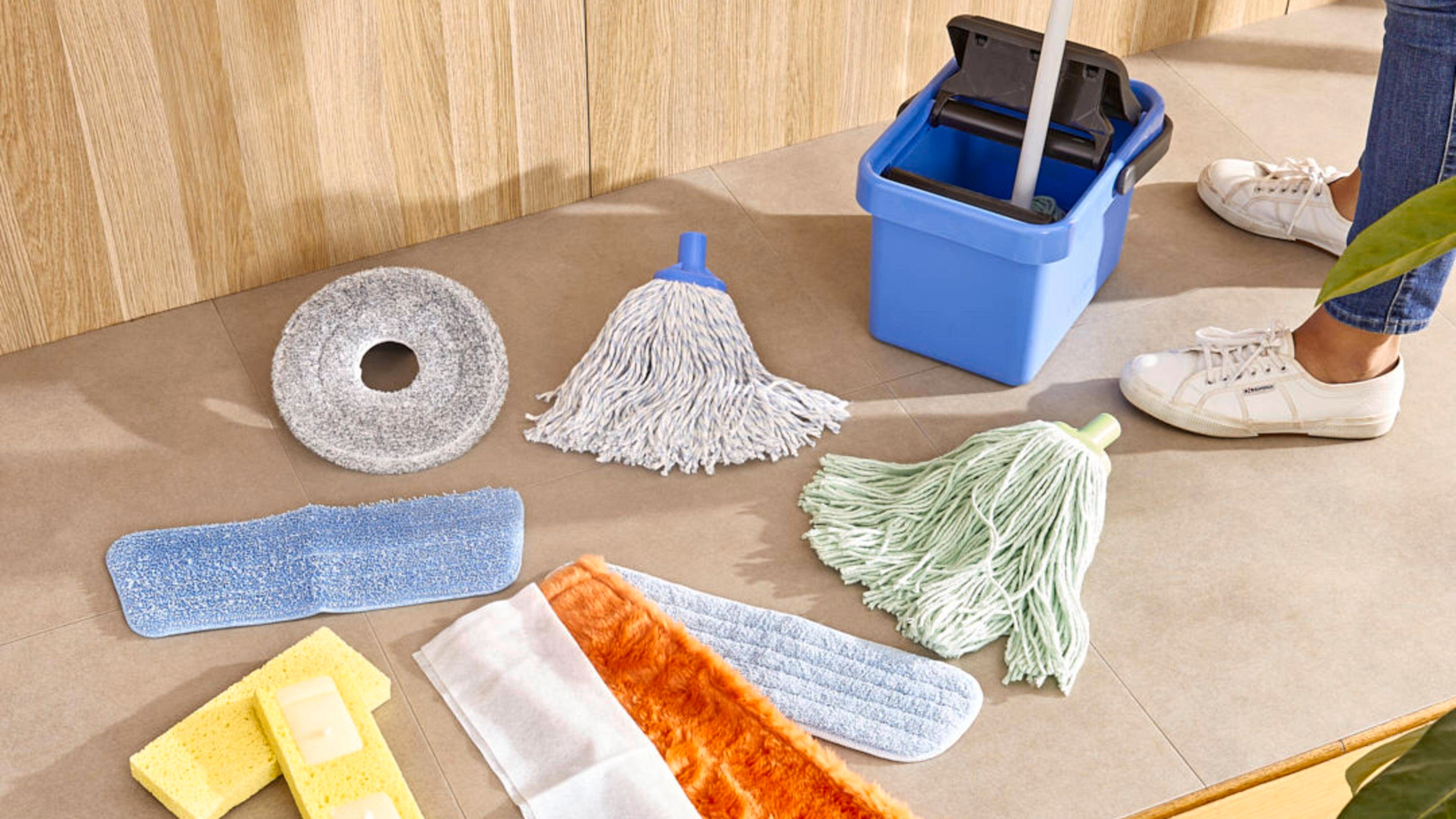 Selection of commercial cleaning supplies arranged on a tiled floor, including mop heads, floor pads, sponges, microfiber pads, a cleaning bucket, and dusting accessories.