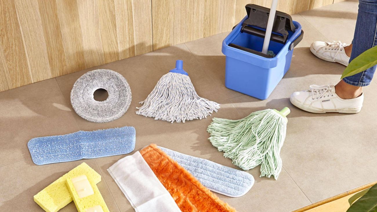 Selection of commercial cleaning supplies arranged on a tiled floor, including mop heads, floor pads, sponges, microfiber pads, a cleaning bucket, and dusting accessories.
