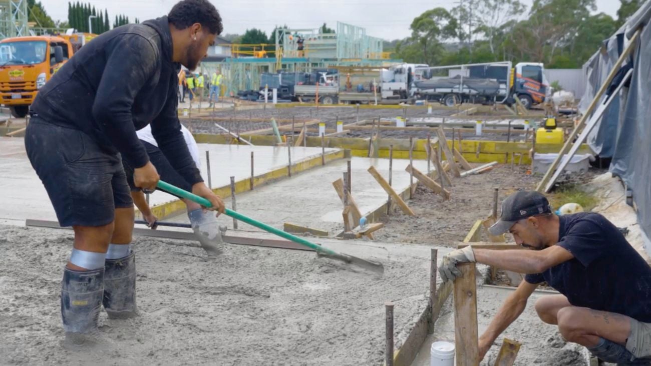 Two workers shape wet concrete beside timber formwork; one pulls a screed with a green handle, the other braces a timber peg. Part‑built slabs, framing, and trucks are visible in the background on a construction site.