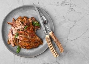 Carved lamb spit roast garnished with rosemary on a grey platter with a serving fork and knife.