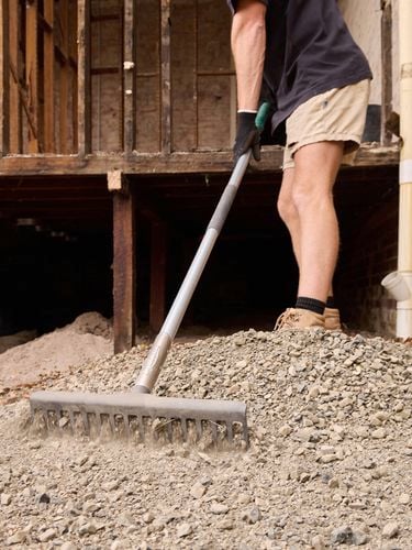 Person using a rake to spread gravel near a wooden structure and brick wall with a drainage pipe.