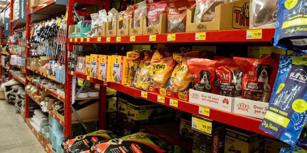 Bunnings pet supplies section featuring dog treats, pet food, leads, collars and accessories displayed on red warehouse shelving. Packaged pet products line the shelves, with large bags of dog food stored on the lower level and category signage visible overhead.