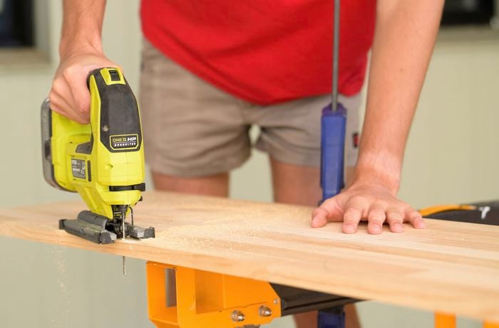 A person is using a jigsaw to carefully cut along the curves on a wooden board that is clamped to a workbench.