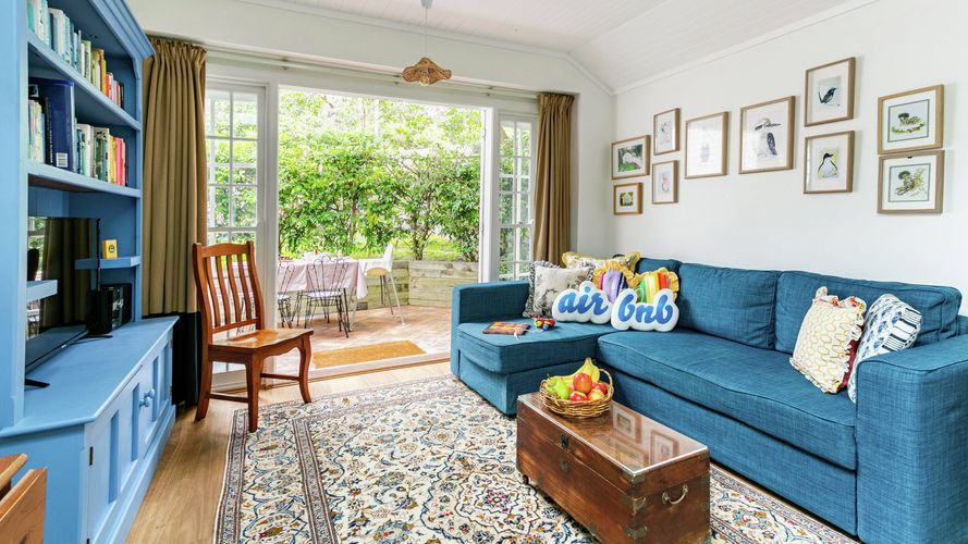 Welcoming living room featuring a blue sectional sofa, built-in blue shelving, patterned rug and timber coffee table, with French doors opening onto a leafy outdoor patio.