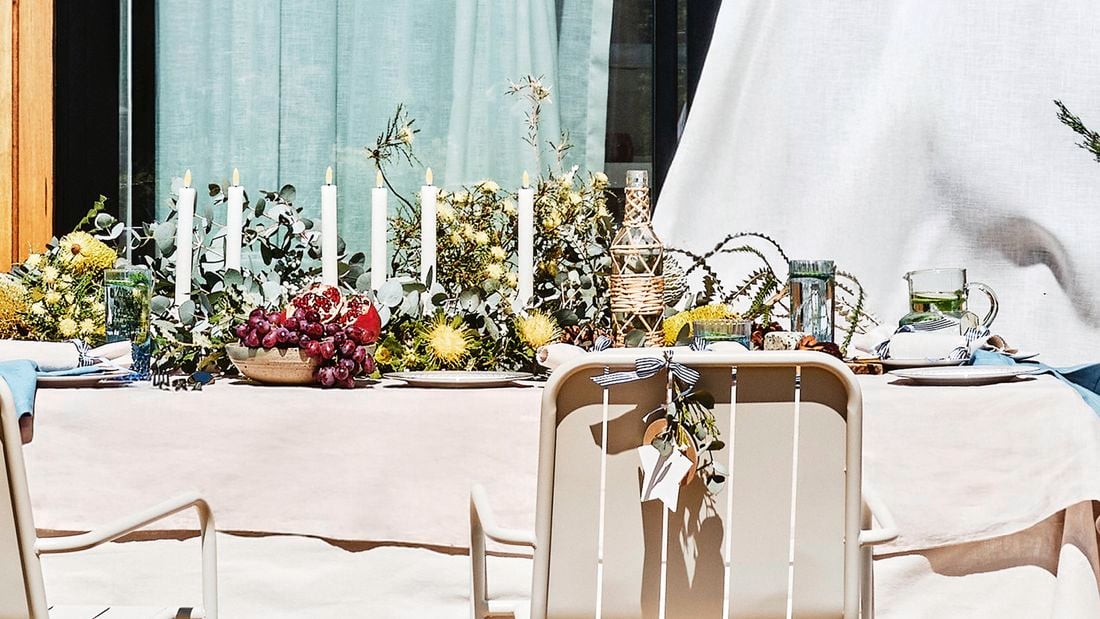 Outdoor table setting with a light tablecloth, a long greenery centrepiece holding white taper candles, and bowls of grapes and other fruit. Sliding glass door with pale curtains behind; beige chairs, one tied with a ribbon and foliage, and a twine‑wrapped bottle on the table