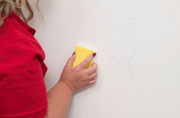 A person in a red T shirt is removing pencil marks on a white wall using a magic erase