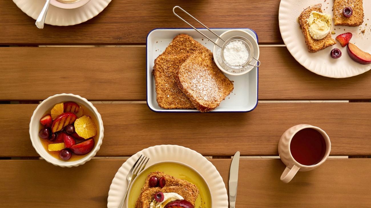 A breakfast setting set shot from above on a wooden table with French toast topped with fruit and yoghurt, a bowl of mixed fruit salad, a teapot, cup of tea and a bowl of yoghurt.