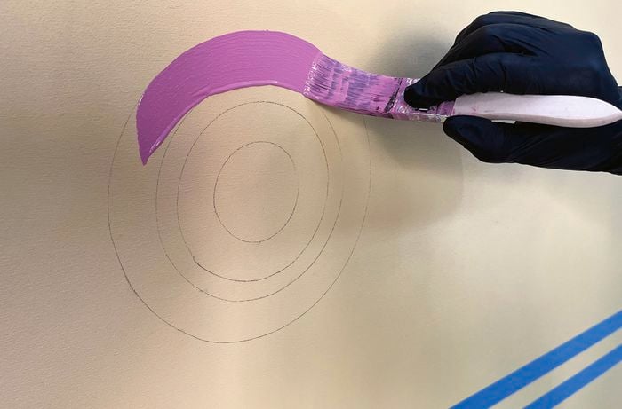A gloved hand paints a purple circle onto a yellow-painted timber surface with a paintbrush.