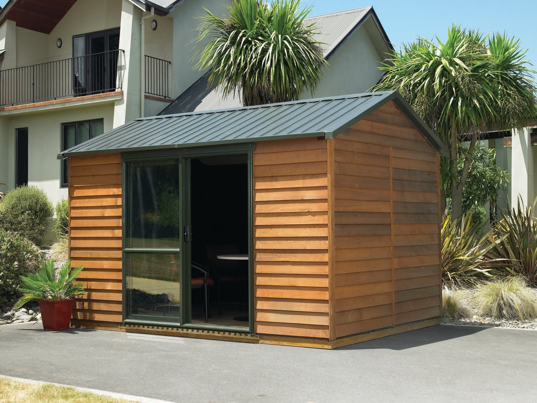 A backyard shed made from stained weatherboards with a fern in a pot plant outside