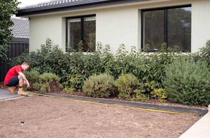 A person in a red shirt is measuring a garden area with a tape measure, preparing the ground for turf installation.