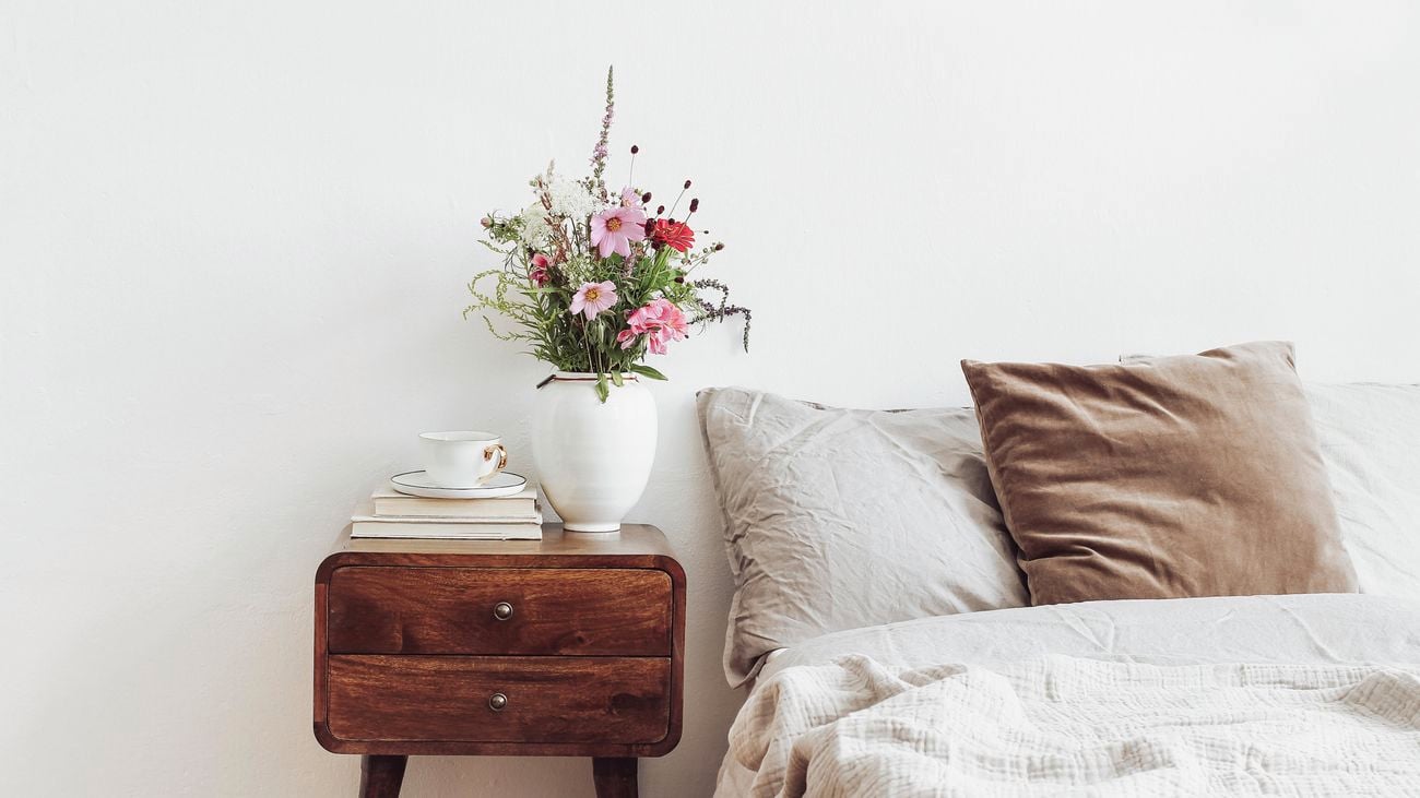 A cup, a vase of flowers, and books on a retro wooden bedside table next to a bed with neutral-coloured linens and cushions.