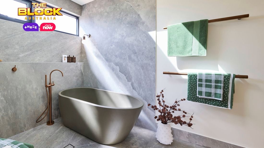 Modern bathroom with matte gray freestanding tub, stone-look gray tiles, wall-mounted faucet, green towels on wooden racks, and natural light from a high window. 