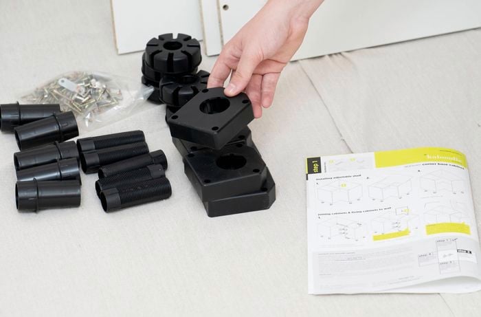 A person is assembling a corner base cabinet, with various black plastic components, screws, and an instruction manual laid out on a light-colored surface