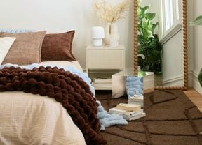 A bedroom with a mirror cube on a brown rug next to a bedside table with a lamp and vase, and a bed with brown and blue blankets. A tall, beaded floor mirror and a large green plant are also visible.