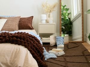A bedroom with a mirror cube on a brown rug next to a bedside table with a lamp and vase, and a bed with brown and blue blankets. A tall, beaded floor mirror and a large green plant are also visible.