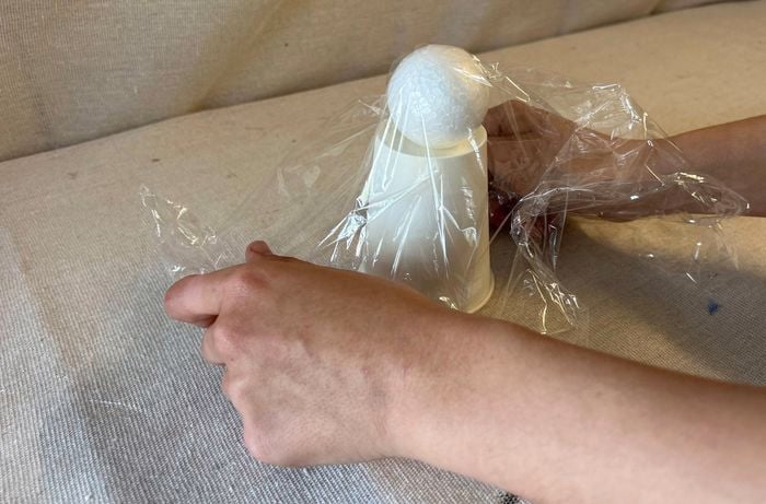 A pair of hands drape plastic wrap over a styrofoam ball on an upturned paper cup.