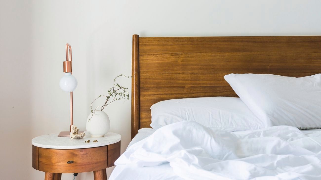A bed with white sheets and wooden headboard with a bedside table next to it with a lamp and plant decorations