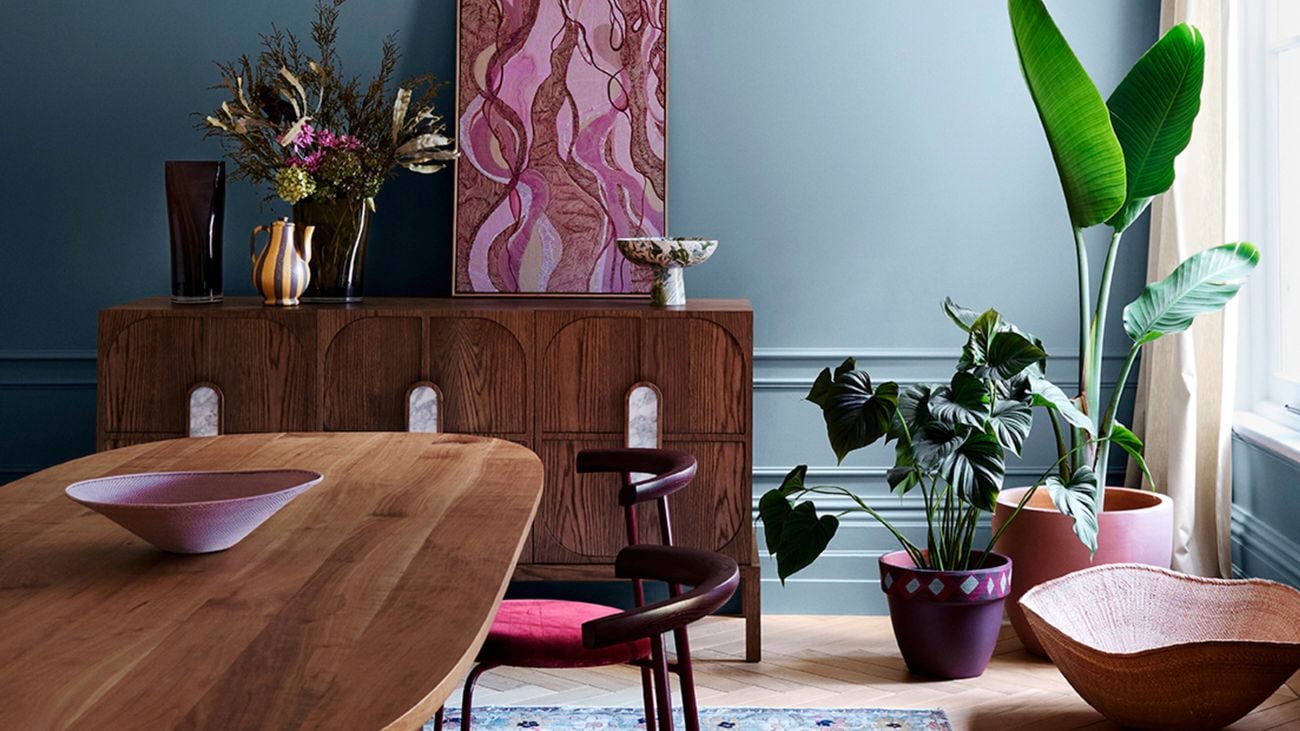 A dining room with a wooden table and chairs on top of a rug, with a side cabinet and pot plants in it.