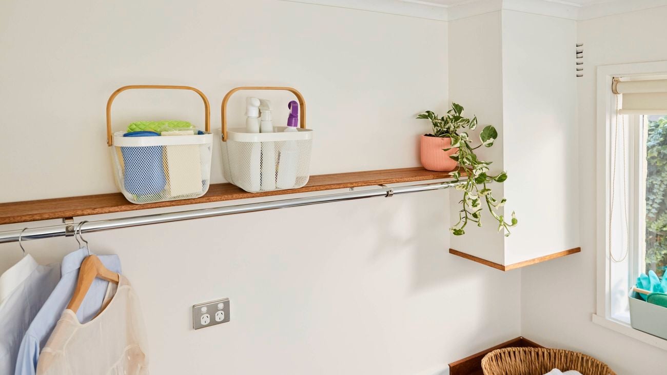 Wall-mounted wooden shelf with two metal baskets holding cleaning supplies, a hanging rail with shirts below, and a trailing potted plant near a window.