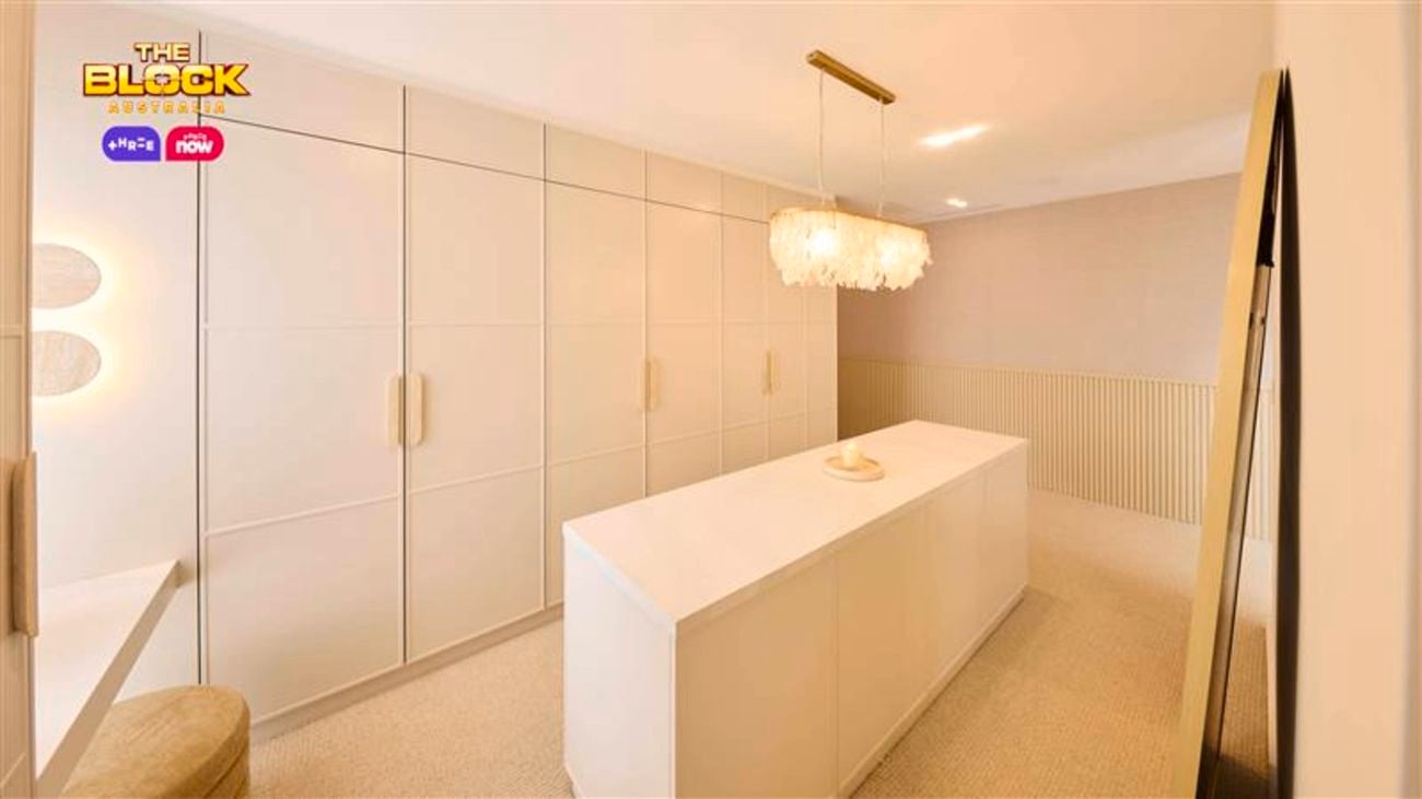 A modern walk-in wardrobe featuring full-height white cabinetry with large handles, a central white island bench, a decorative chandelier above, and neutral-toned walls and flooring.