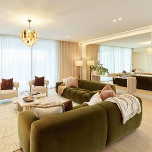 Contemporary living room featuring curved olive-green sofas, neutral armchairs, a chandelier and soft floor-to-ceiling curtains.