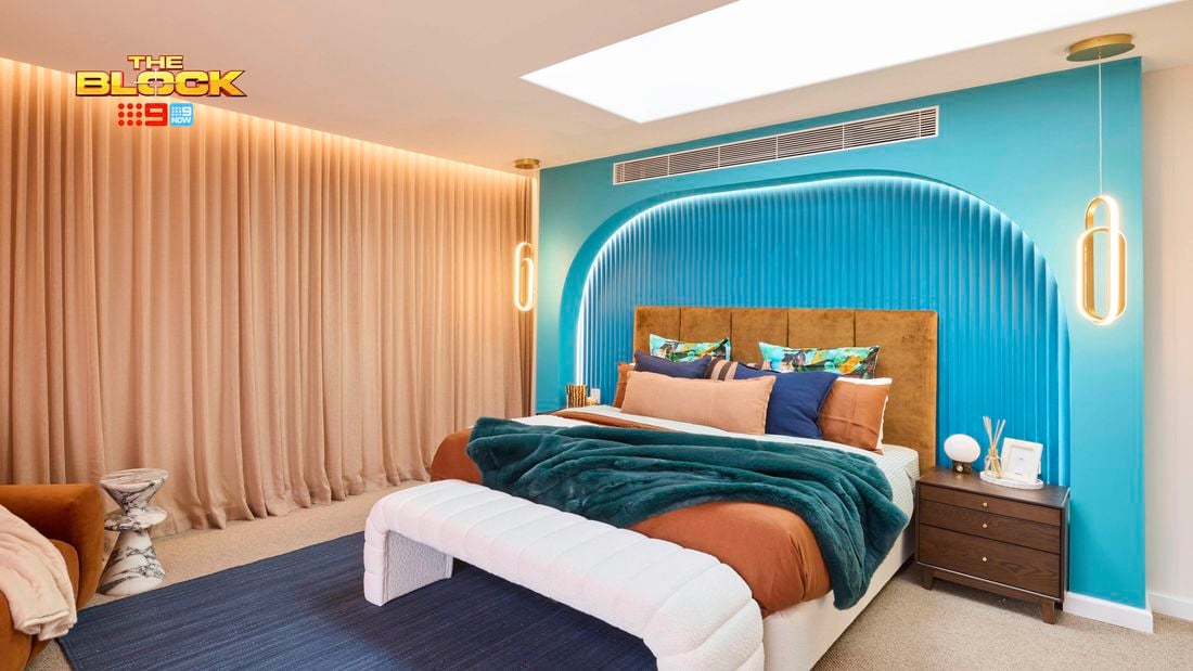 A modern bedroom featuring a large bed with white and brown bedding, colorful pillows, and a green throw blanket. The blue headboard wall has an arched design and pendant lights on either side. A wooden nightstand with decorative items sits next to the bed. The room includes a blue rug, beige curtains, and a rectangular skylight in the ceiling. 