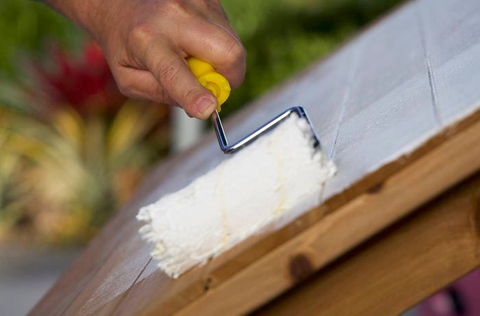 A hand using a mini roller to apply white paint to a cubby house exterio