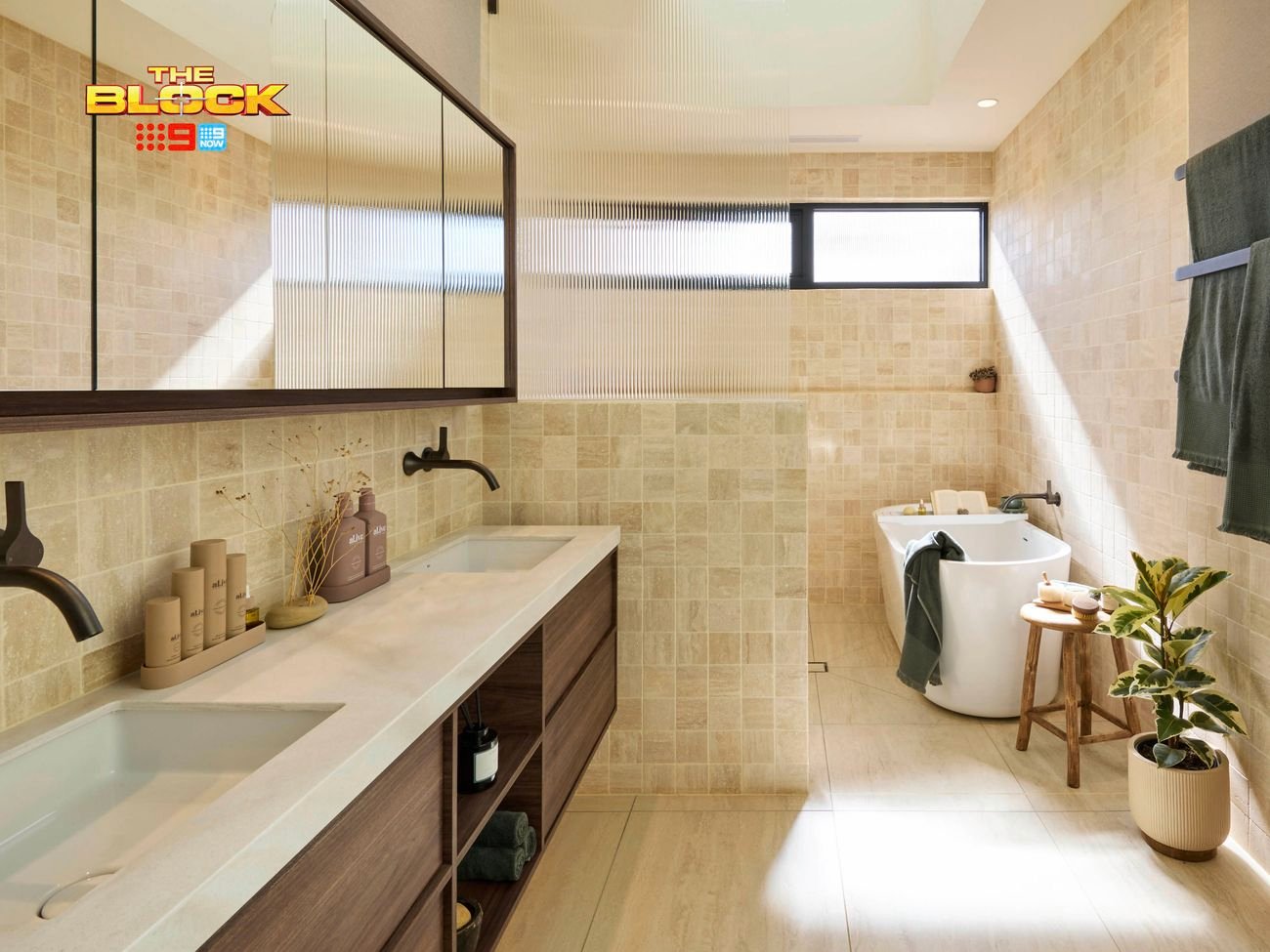 Modern bathroom with double sink vanity, black faucets, freestanding tub, beige tiles, green towels, and natural light from a window. 