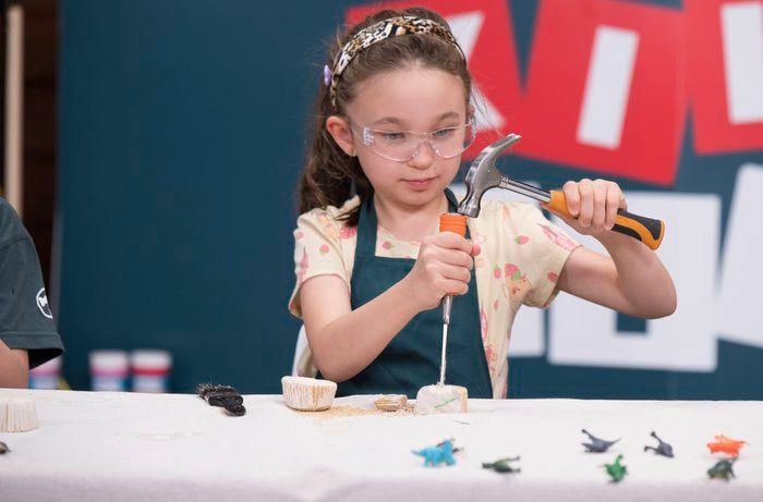 A young girl wearing safety goggles and an apron is using a hammer and chisel to break open a plaster block to reveal the fossil.