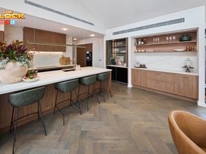 A modern white kitchen with brown cupboards, bar stools at the benchtop with a vase with flowers in it and two bowls with items in them on it.