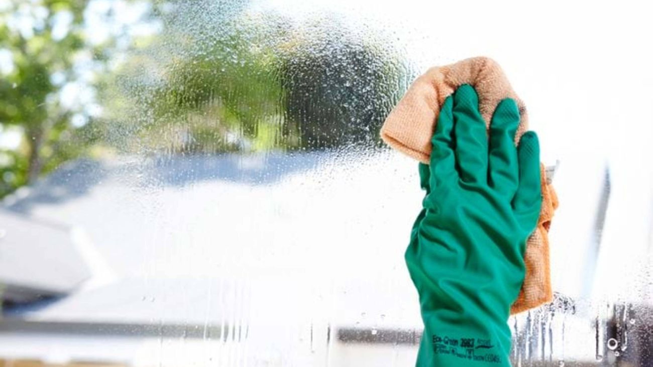 Gloved hand using a cloth to clean a large glass window covered in cleaning solution, with an outdoor view blurred in the background.