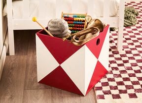 Red-and-white geometric storage box in a child’s room holding toys on a burgundy-and-cream chequered rug beside a white bed.