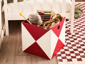 Red-and-white geometric storage box in a child’s room holding toys on a burgundy-and-cream chequered rug beside a white bed.