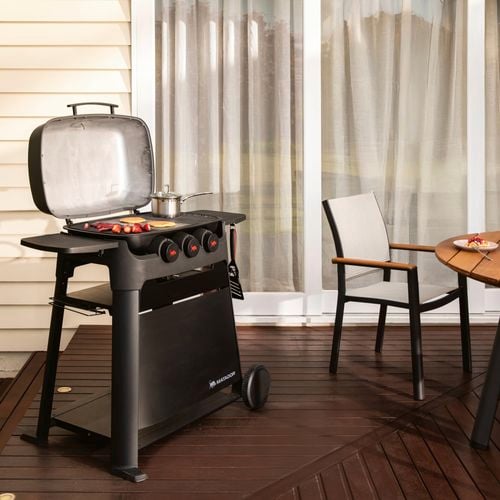 Outdoor barbecue grill on a wooden deck with food cooking, next to a round table set for a meal