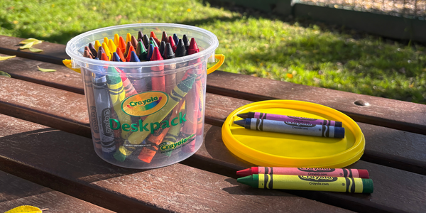 Container of Crayola coloured crayons with selected crayons arranged beside the lid on a wooden outdoor table.
