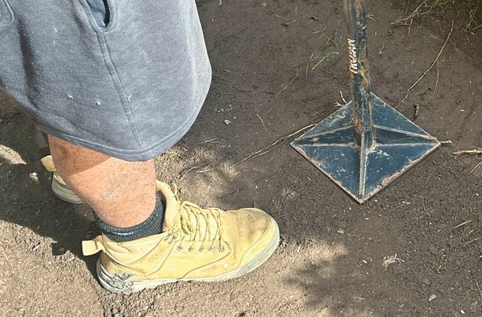 A person wearing work boots and in shorts uses a tamper to compact soil