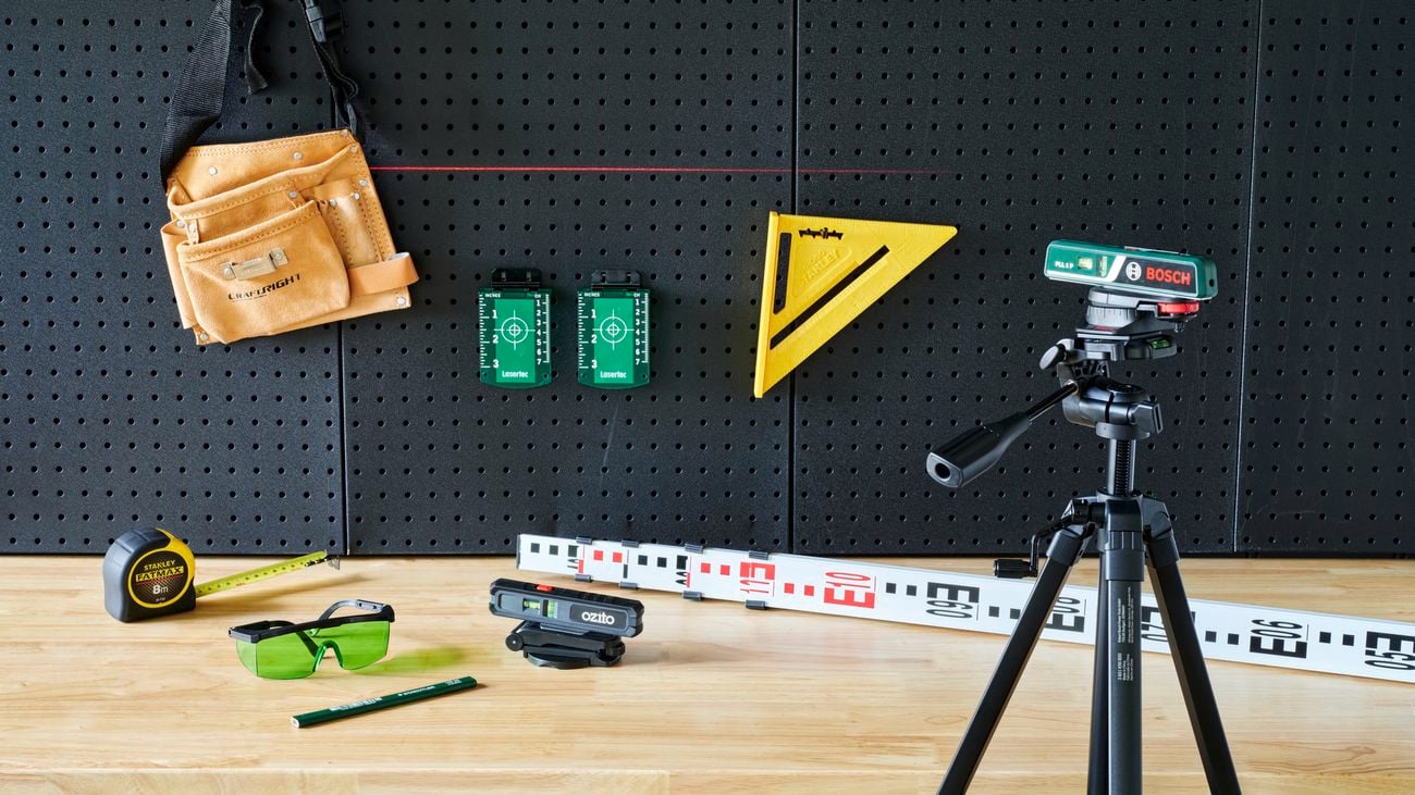 Bosch laser level on tripod pointing onto black wall with accessories on timber bench including tape measure.