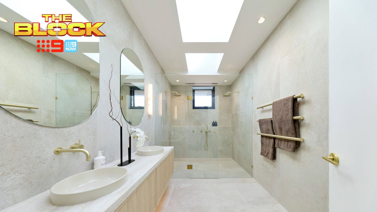Modern bathroom with a long timber vanity, double white basins, round mirrors, brass tapware, walk-in shower and neutral stone finishes. “The Block” logo appears in the upper-left corner.