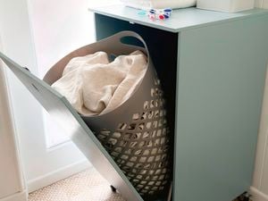 A light blue tilt-door laundry hamper cabinet with a grey hamper inside, holding white laundry.