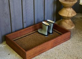 Brown wooden tray with gumboots on it, on the ground, with a fence behind it.