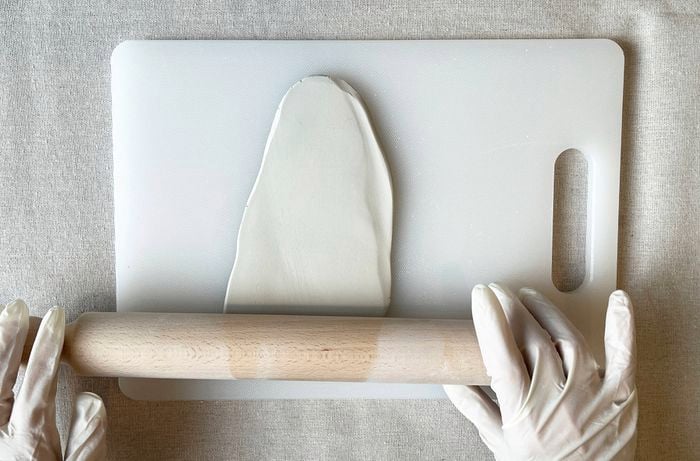 A pair of hands wearing white gloves rolls out white clay using a wooden rolling pin on a white cutting board.