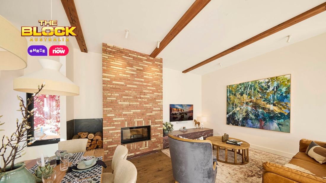 A modern living room featuring a central brick fireplace, exposed wooden ceiling beams, and a large abstract painting on the right wall. Furnished with a brown leather sofa, two gray armchairs, and a round wooden coffee table atop an area rug. A dining table set for a meal is visible to the left.
