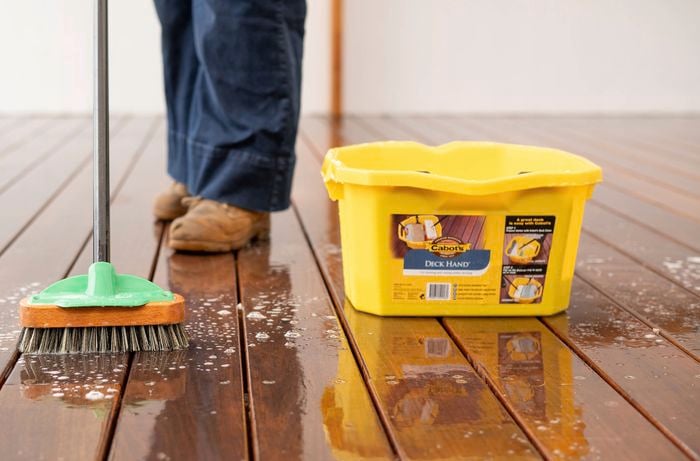 A Bunnings Team Member is scrubbing a wooden deck with a broom and a yellow bucket nearby.