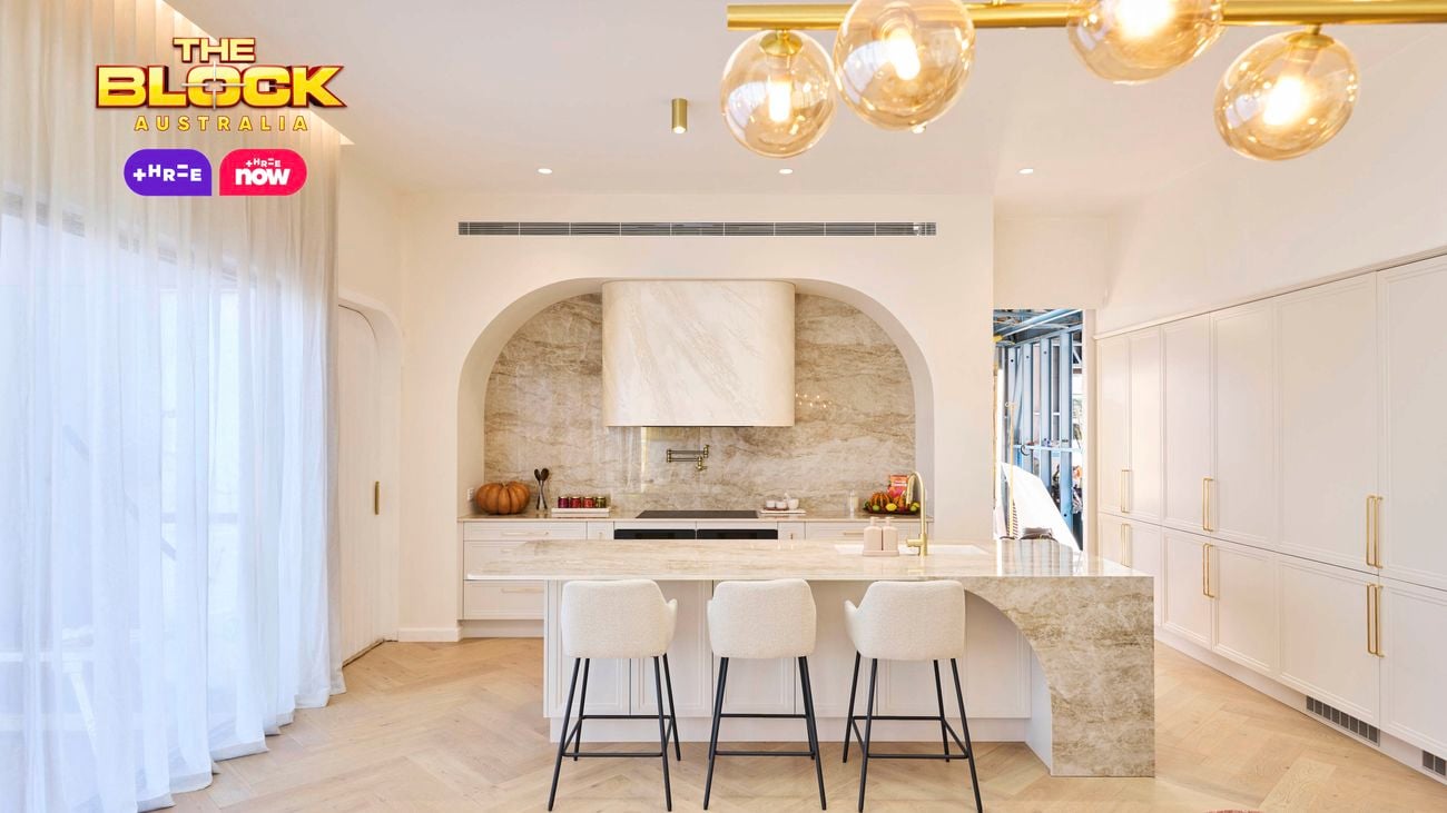 Bright, modern kitchen from The Block, featuring a marble island with four bar stools, arched range hood with stone backsplash, white cabinetry, gold fixtures, and a contemporary glass bubble chandelier.