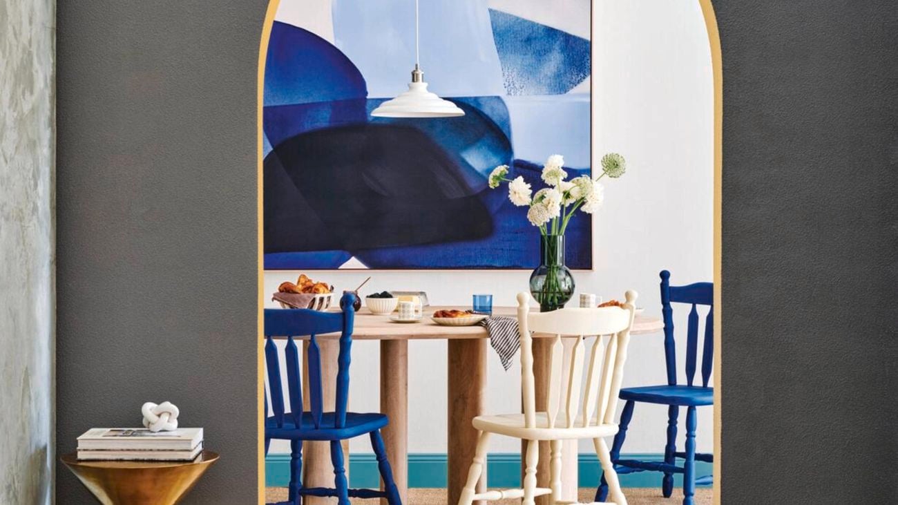 Grey wall with arched doorway, opening out into a white room with a blue abstract painting and a wooden dining table with blue and white chairs.