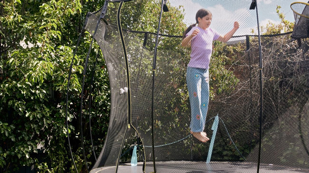 Young girl in purple shirt and light blue pants mid-jump on a backyard trampoline surrounded by safety netting and lush green trees on a sunny day