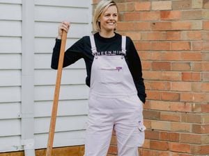 Person wearing light-colored overalls, a black long-sleeve shirt with visible text “GREEN HIP,” and brown boots, standing on grass and holding a garden spade upright near a white weatherboard wall and brick wall.