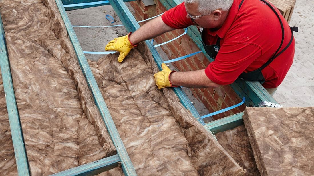 A person wearing protective gear placing an insulation batt between floor joists
