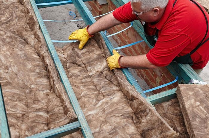 A person wearing protective gear placing an insulation batt between floor joists